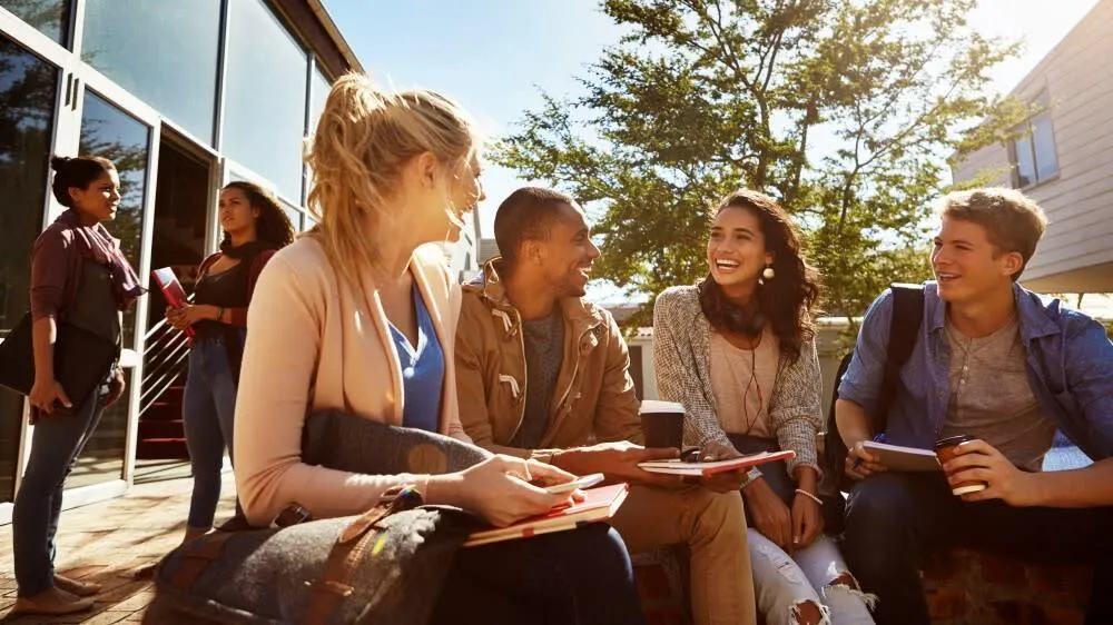 Image: A diverse group of college students engaging in conversation and laughter on a campus lawn, showcasing the joy and camaraderie of making friends at college in the United States. The image is associated with the blog post 'How to Make Friends at College in the US - By Esposearch.